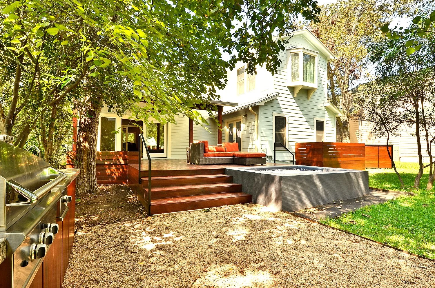 Backyard patio area with a raised wooden deck, outdoor sofa, grill, and surrounding trees and grass.