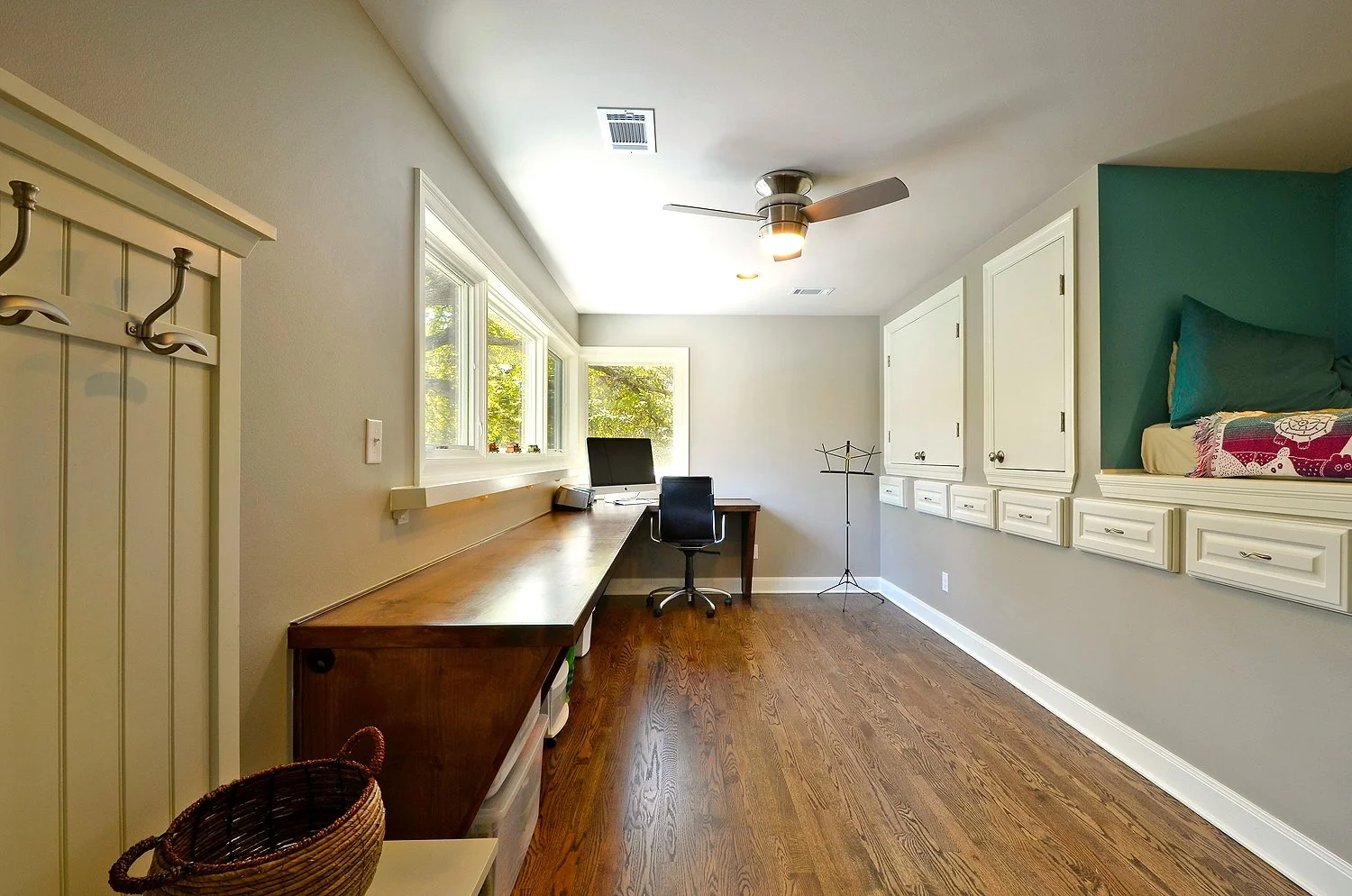 Home office with a wooden desk, black office chair, and a computer monitor near three large windows, with built-in white cabinets on the right and a small nook with bedding above. Wooden flooring throughout, ceiling fan with light, and natural light 