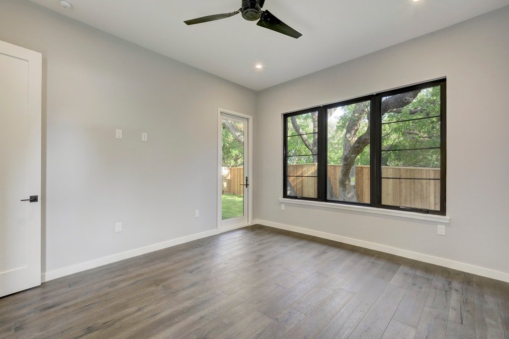 Empty room with large black-framed window showing trees outside, a white door with glass panel leading to the backyard, light gray walls, wood laminate flooring, ceiling fan, and recessed ceiling light.