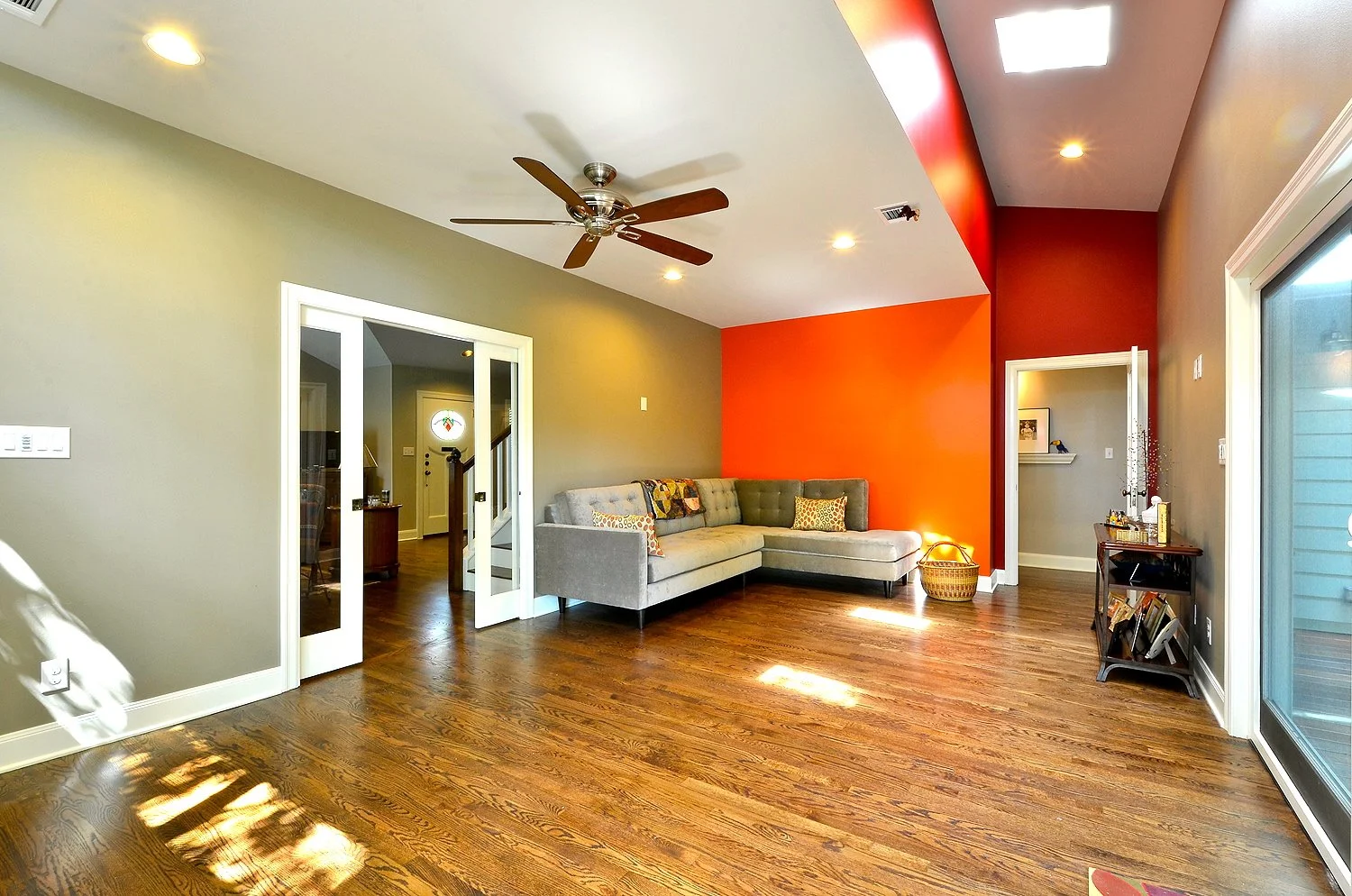 Living room with hardwood floors, green and red walls, a ceiling fan, a grey sectional sofa with decorative pillows, a sliding glass door, and a small side table with books and decorative items.