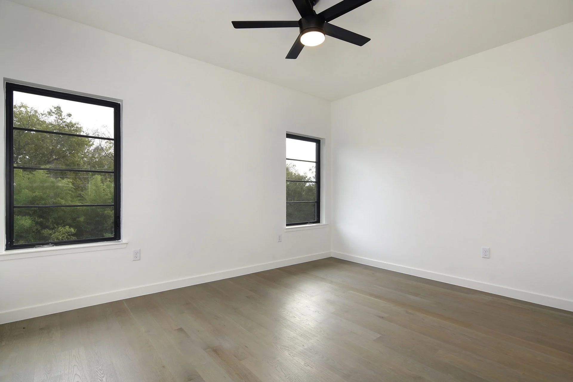 Empty white room with two windows, ceiling fan, light-colored wooden floor, and white electrical outlets.