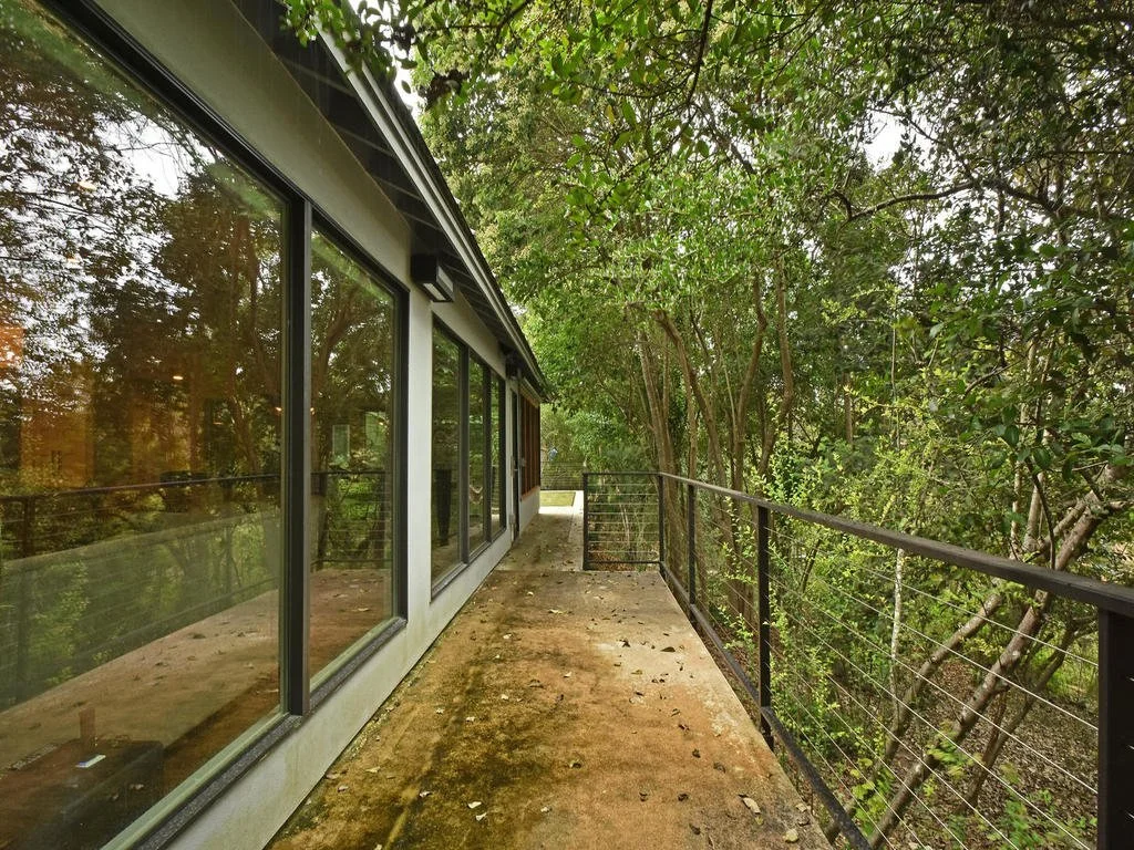 A balcony with black metal railings extends from a house with large glass windows, surrounded by green leafy trees.