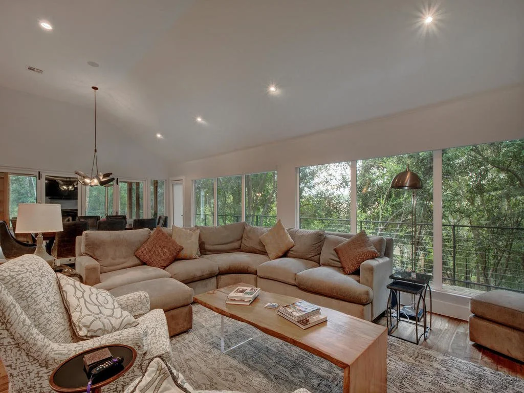  Spacious living room with large beige sectional sofa, patterned armchair, coffee table with magazines, large windows showing green trees outside, and a modern chandelier.