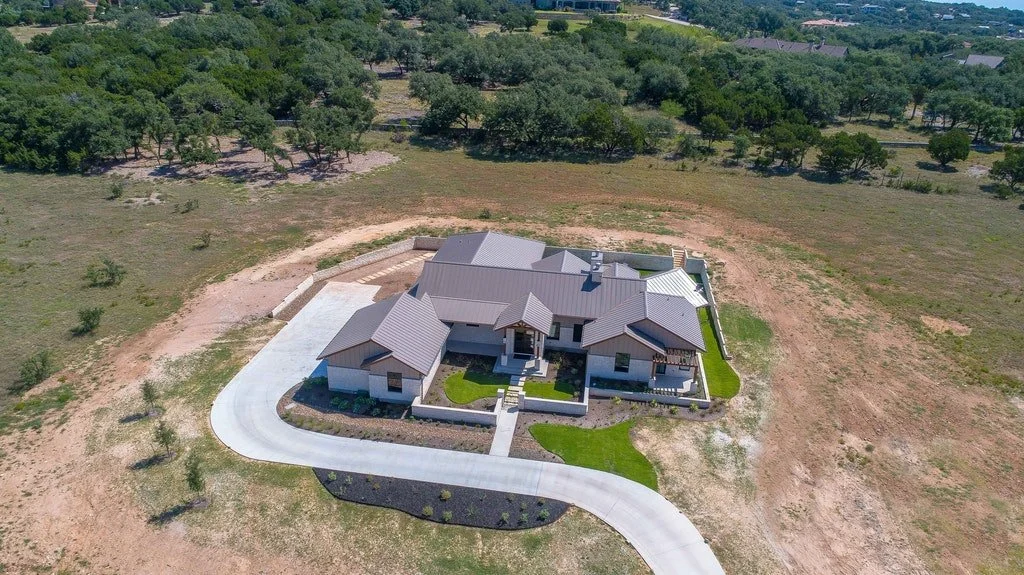 A large, modern house with gray roofing and white walls, surrounded by a landscaped yard and a curved driveway, located in a semi-rural area with trees and open land.