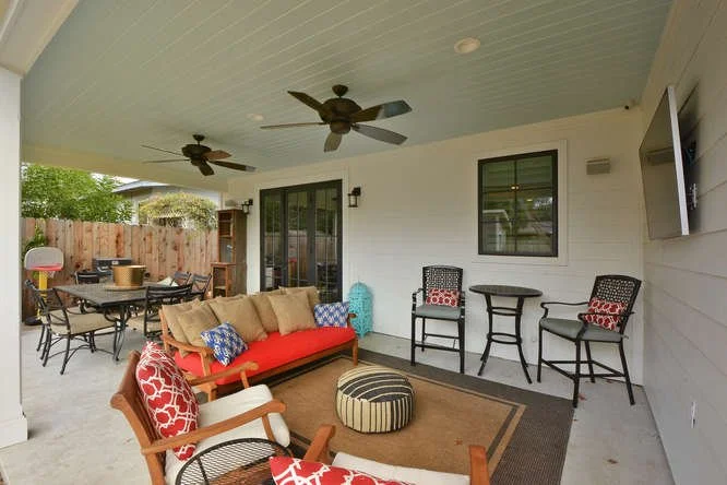 Covered outdoor patio with seating area, ceiling fans, and a mounted TV, featuring a colorful sofa, chairs, and a dining table.