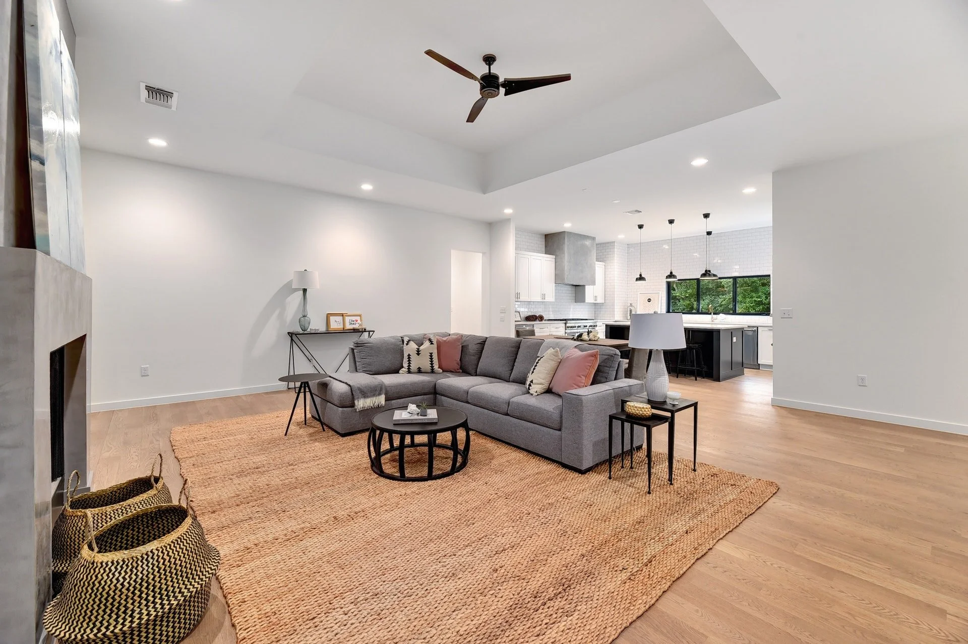 Open-plan living room with gray sectional sofa, black coffee tables, and a woven area rug, adjacent to a kitchen with white cabinets and black accents, large window with green trees outside, ceiling fan, and white walls.