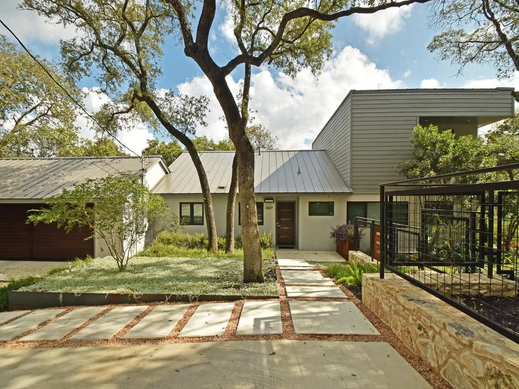 Modern house with a metal roof, trees, a small front yard, a concrete pathway, and a black metal fence.