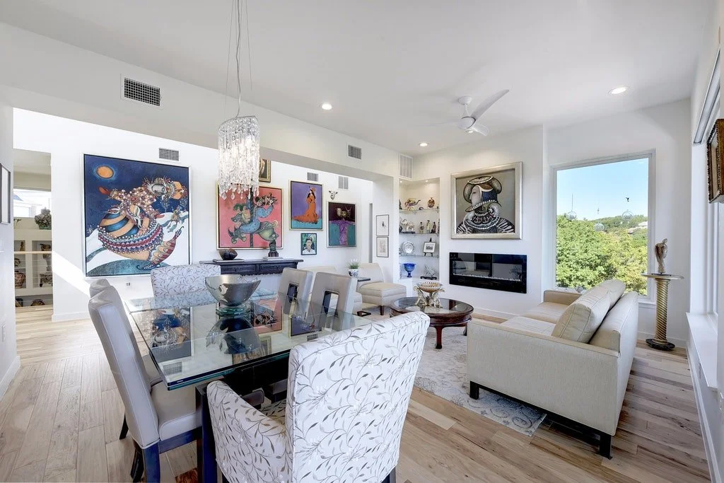 Bright and modern living room with white walls, hardwood floors, large window with a view of green trees, white ceiling fan, and contemporary furniture including a white sofa, armchair, and glass dining table with upholstered chairs. Decor includes c