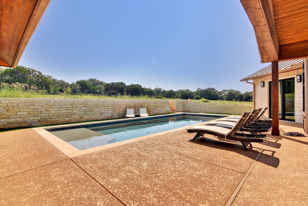 Outdoor patio with a small swimming pool, four lounge chairs, and two white chairs facing the pool, overlooking a grassy landscape with trees under a clear blue sky.