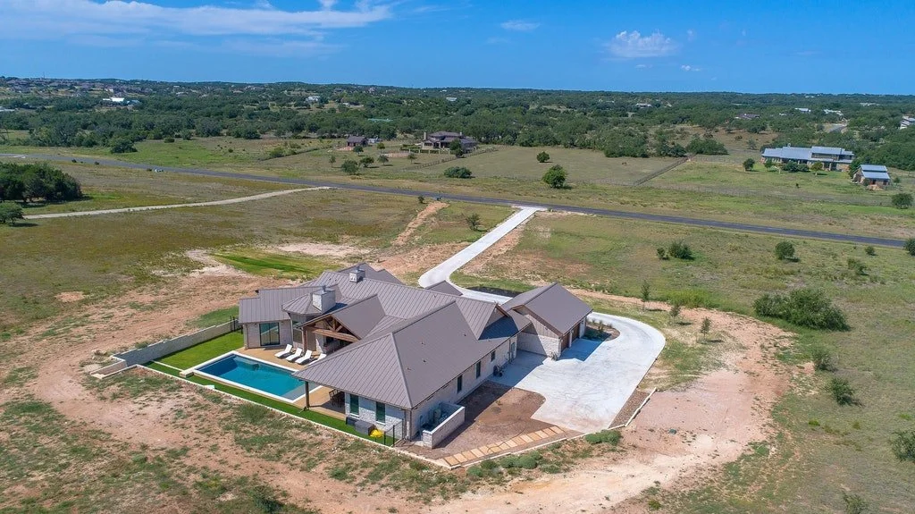 A large house with a gray metal roof, a backyard swimming pool, and surrounding landscape with a driveway leading to a paved road.