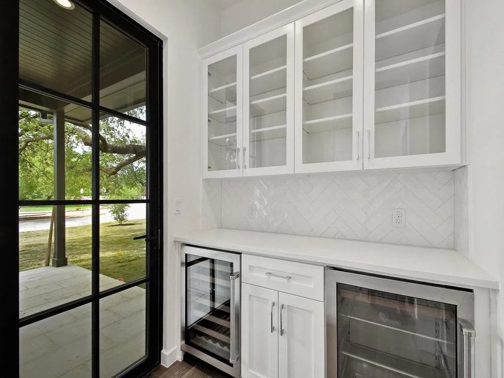 White kitchen with glass-front cabinets, built-in wine cooler, small refrigerator, and a door leading to an outdoor space with a porch and greenery.