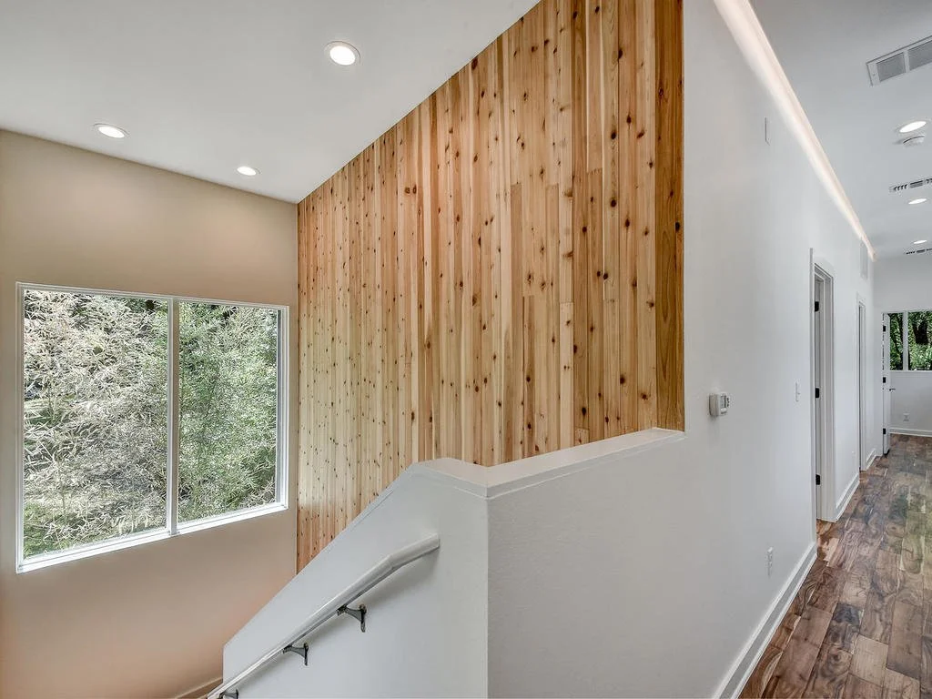Interior view of a home staircase landing with a wooden accent wall, white walls, wood flooring, and large windows showing outdoor greenery.