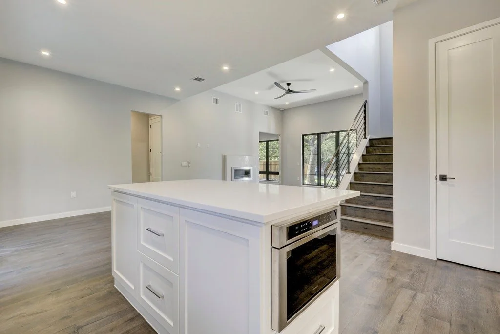 Empty modern kitchen with an island, stainless steel oven, light-colored cabinets, wood flooring, and a staircase leading upstairs.