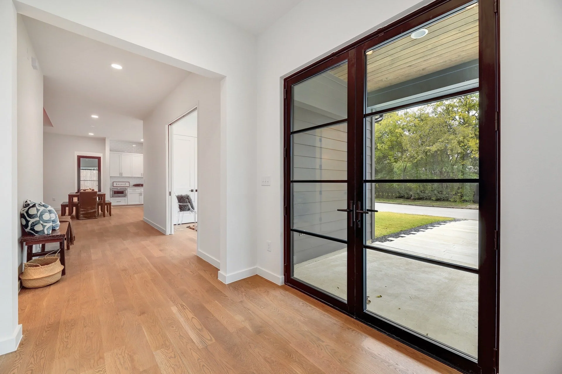 View of a cozy living space with hardwood floors, white walls, and a large glass door leading outside. There is a small sitting area with pillows on a bench, and a glimpse of a dining area and kitchen in the background.