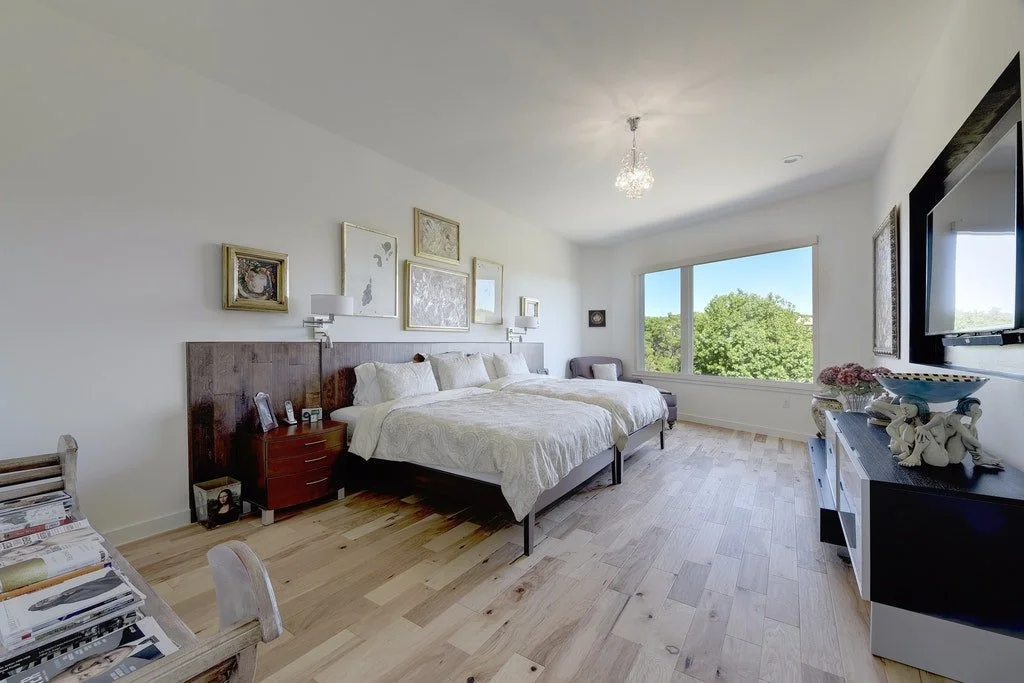 Bright bedroom with large window, white bed, artwork on wall, and wooden flooring.