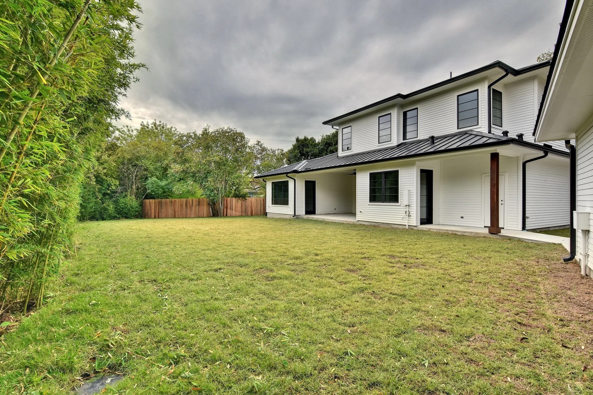 A backyard with a grassy lawn, a white two-story house with black windows, a black metal roof, and a patio. There is a wooden fence, trees, and bushes on the left and in the background, with an overcast sky above.