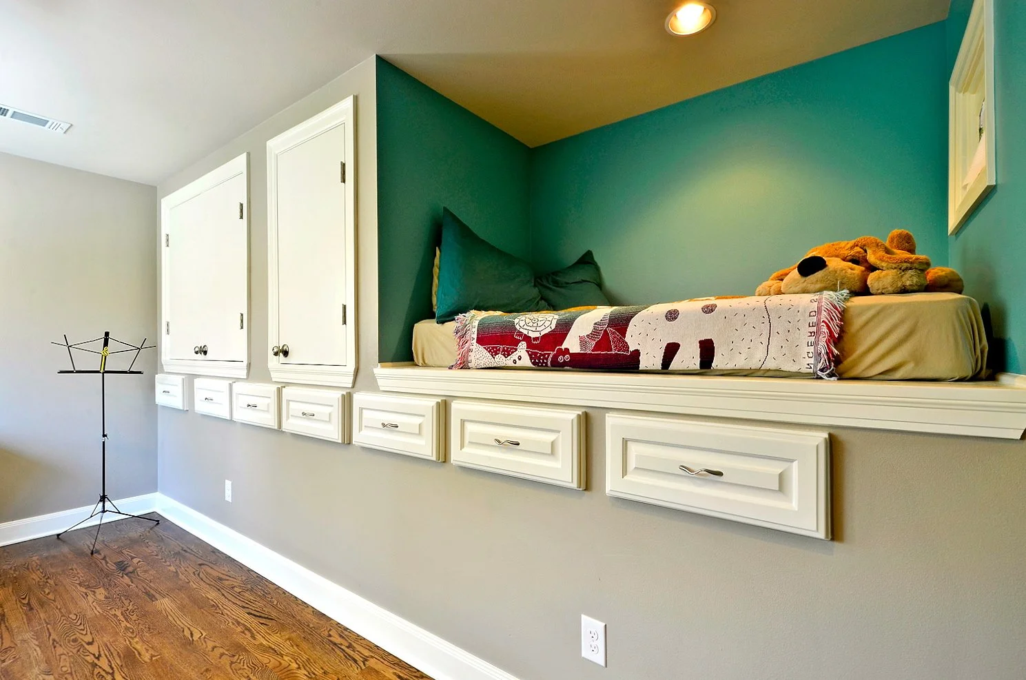 A built-in window seat with a green pillow and a stuffed dog toy, against a teal and beige wall, with storage cabinets below and to the side, hardwood floor, and ceiling light.