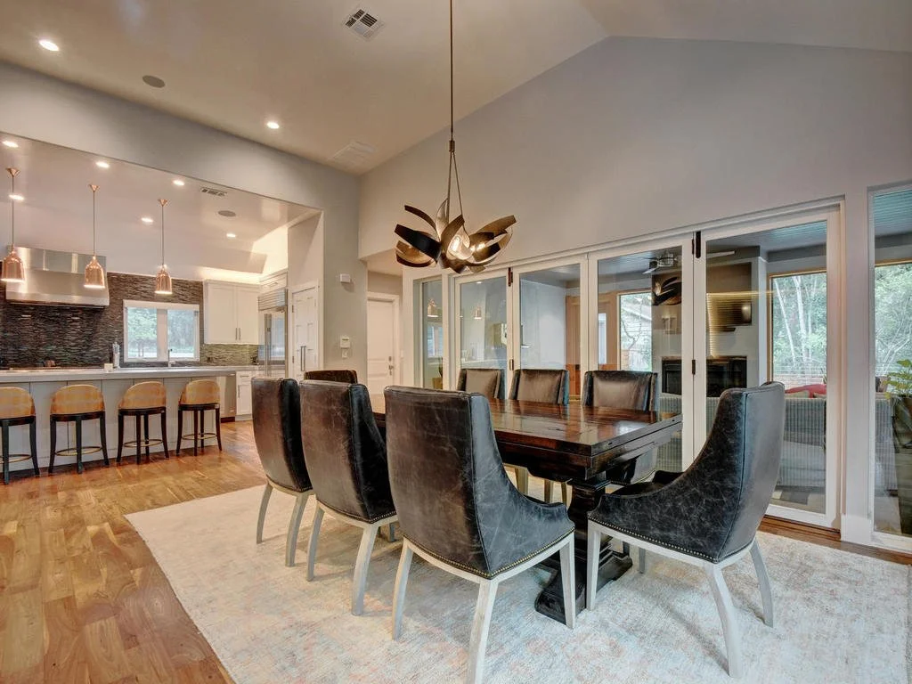 Dining room with a dark wood table, black leather chairs, a modern chandelier, and a large glass sliding door leading to an outdoor area, adjacent to the kitchen with barstools, white cabinets, and pendant lighting.