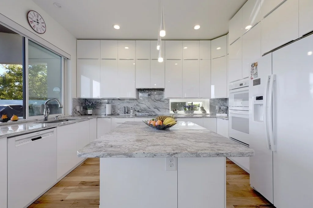 Modern white kitchen with marble countertops and island, large window with trees outside, built-in appliances, and a wall clock.