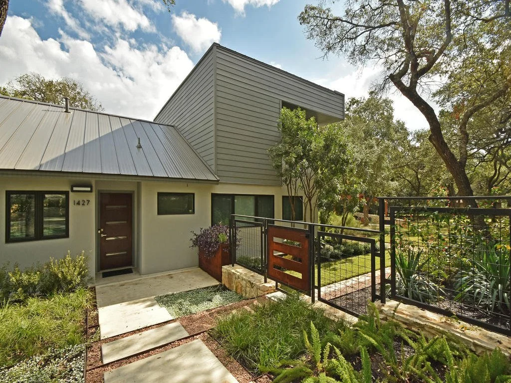Modern house with gray siding, black-framed windows, and a front door, surrounded by greenery and trees, with a pathway and a black metal fence.