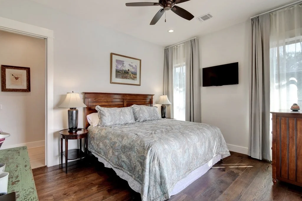 A bedroom with a large bed, wooden headboard, two bedside tables with lamps, a wall-mounted TV, and windows with curtains.