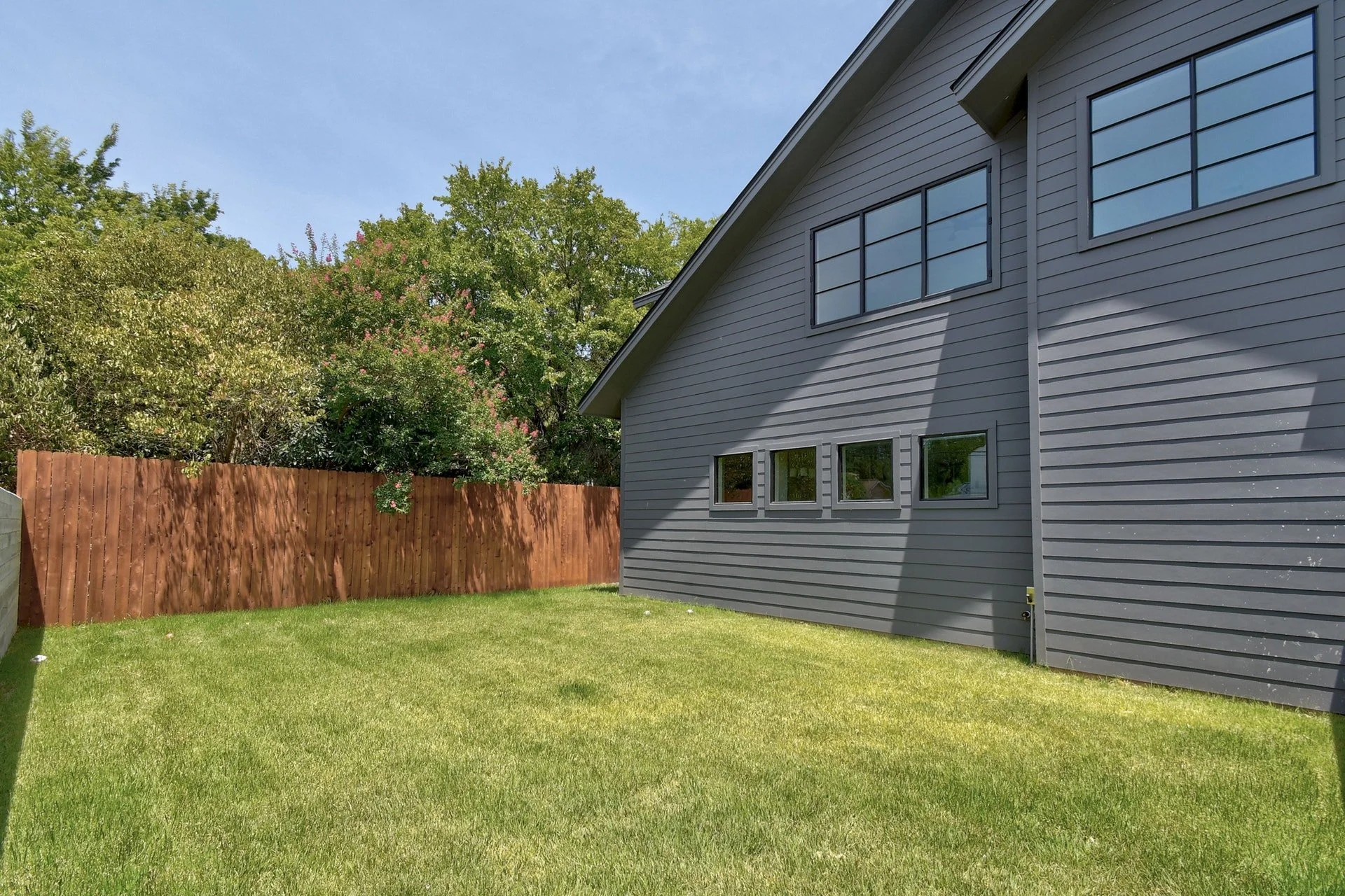 Side view of a gray house with horizontal siding, multiple small windows, a sloped roof, and a wooden fence. There are trees with green leaves in the background and a well-maintained lawn in the foreground.