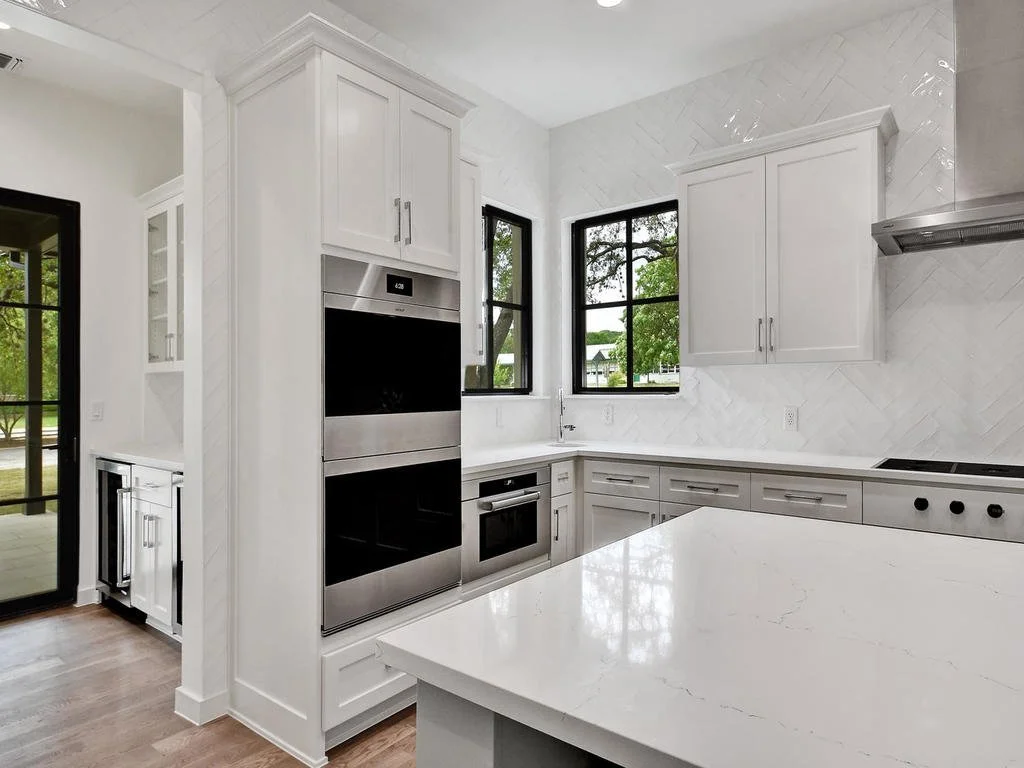 Modern white kitchen with stainless steel double oven, microwave, black window frames, white cabinets, and a marble backsplash.