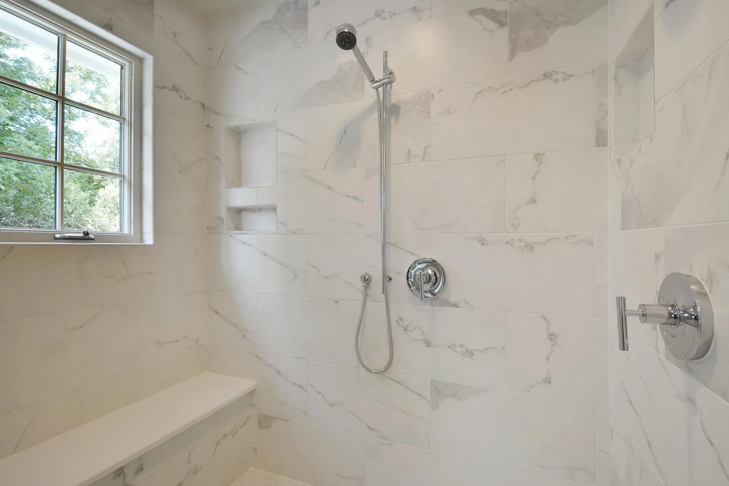 Shower area with a window, marble tiles, a built-in shelf, a handheld shower head, and a wall-mounted shower control