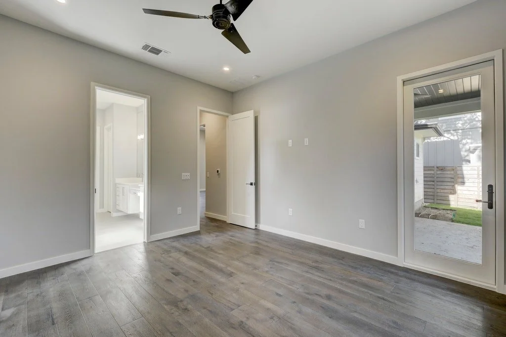 Empty room with gray walls and hardwood flooring, a ceiling fan, a sliding glass door leading outside, and a doorway to a bathroom.