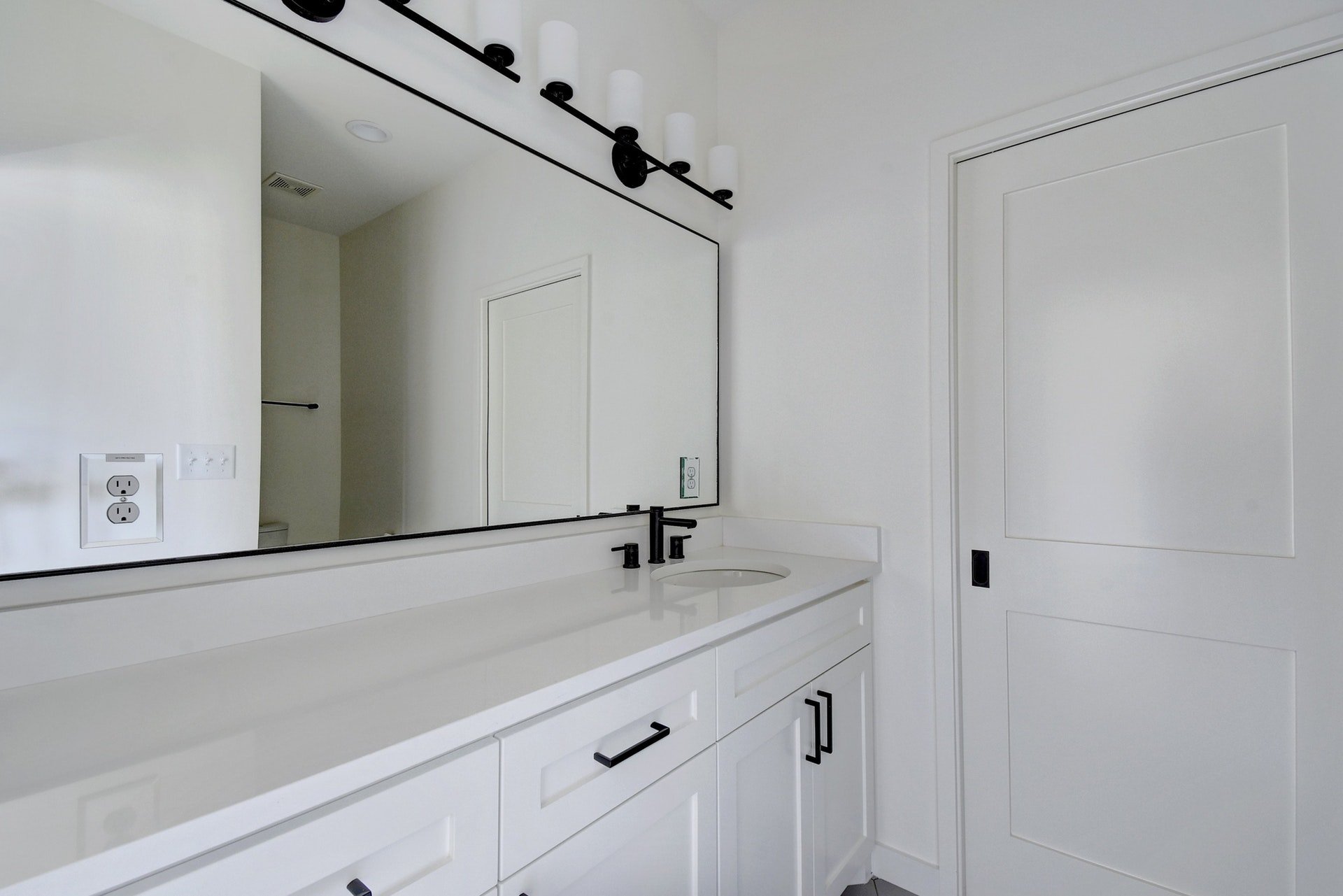 A modern white bathroom vanity with black hardware and a large mirror. A black light fixture with four white cylindrical lights is mounted above the mirror. The countertop has a white sink with a black faucet. There are electrical outlets and switche