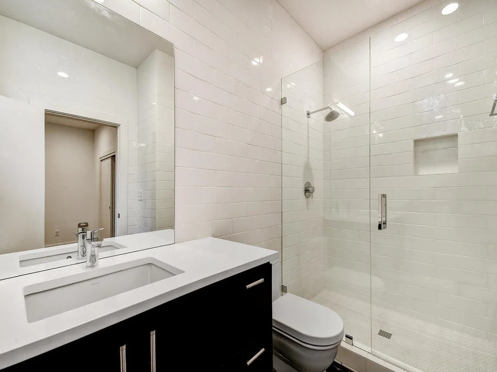 Modern bathroom with white tiled walls, a vanity with a white countertop and a rectangular sink, a large mirror, a walk-in shower with glass door, chrome fixtures, and a separate handheld shower head.