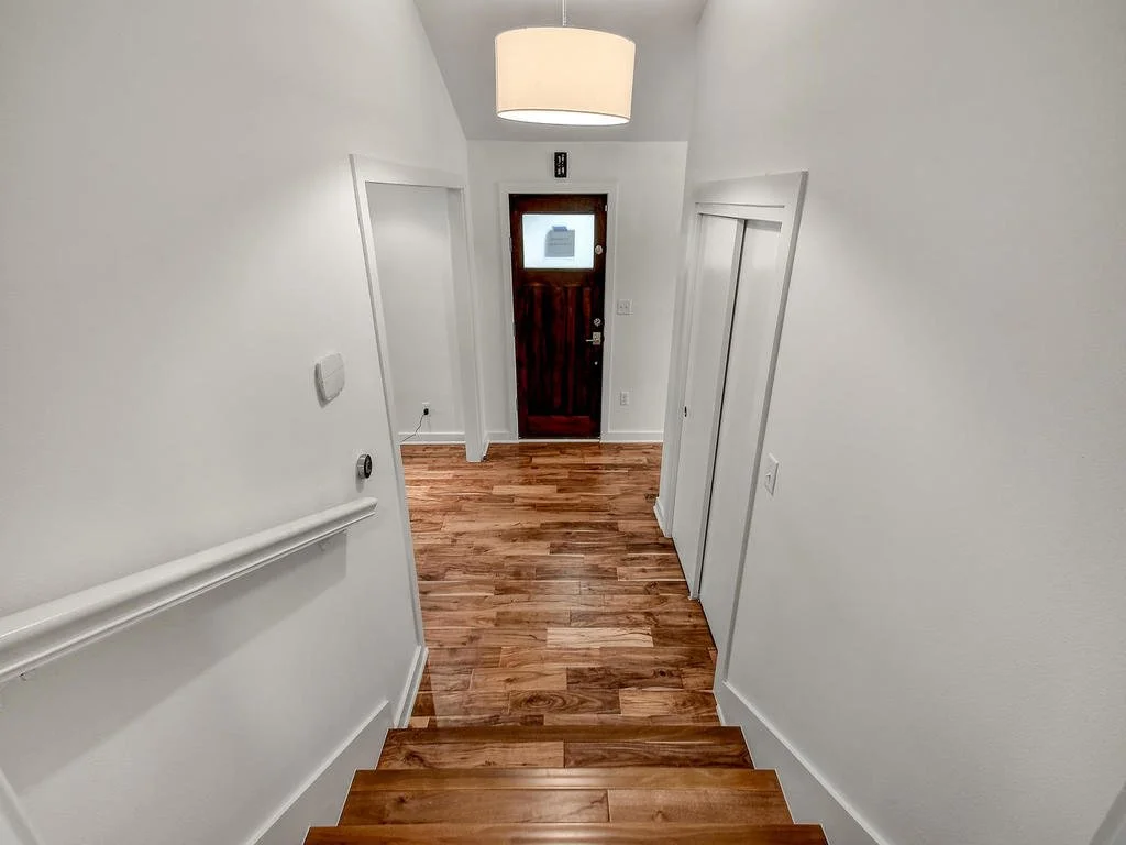 View of a narrow hallway with wooden stairs, wooden front door with a small window, white walls, closet doors, and a ceiling light fixture.