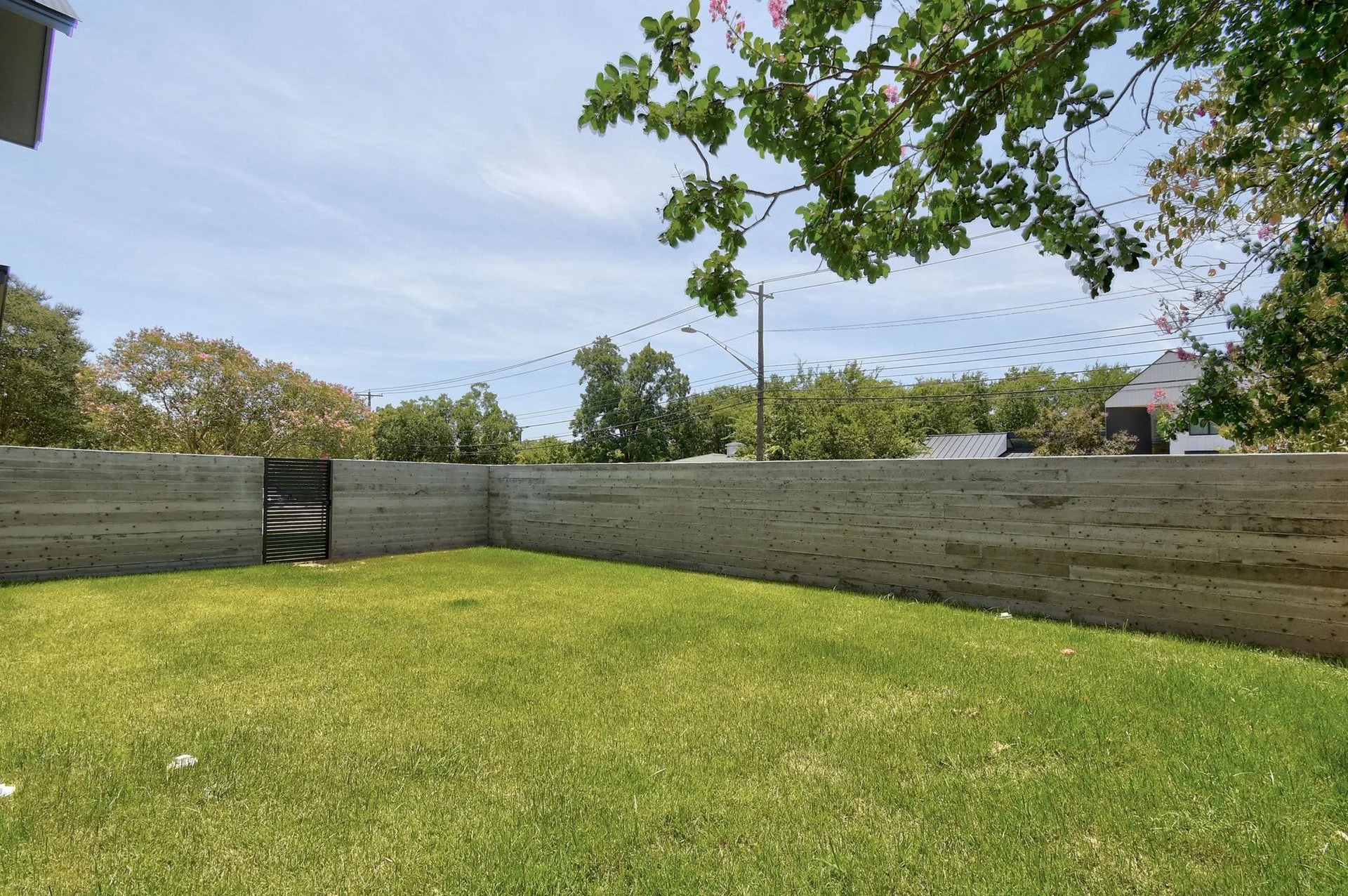 A backyard with a green lawn, surrounded by a gray wooden privacy fence. There are trees with green leaves and pink flowers, and power lines and utility poles are visible in the background. The sky is partly cloudy.