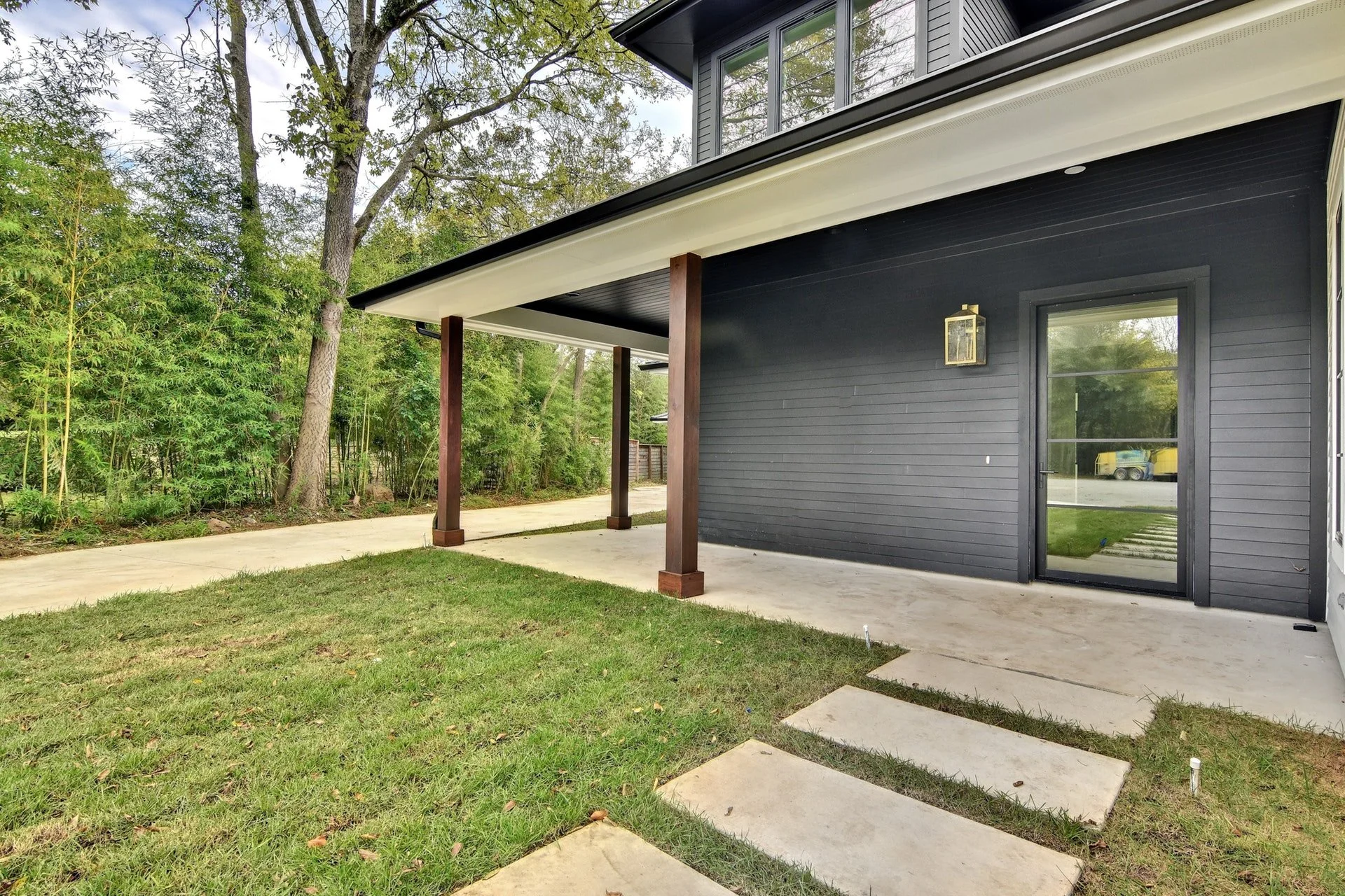 Back patio area of a house with a black exterior wall, a glass sliding door, a gray concrete slab, and wooden support beams for a porch roof. The yard has grass, and there is a wooded area with trees and bushes in the background.