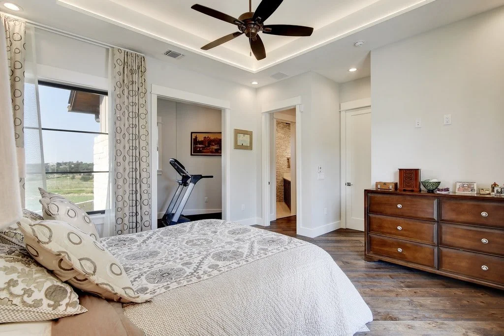 A bedroom with a large bed, decorative pillows, and a view outside the window, with an exercise treadmill near the window, and a wooden dresser on the right side.
