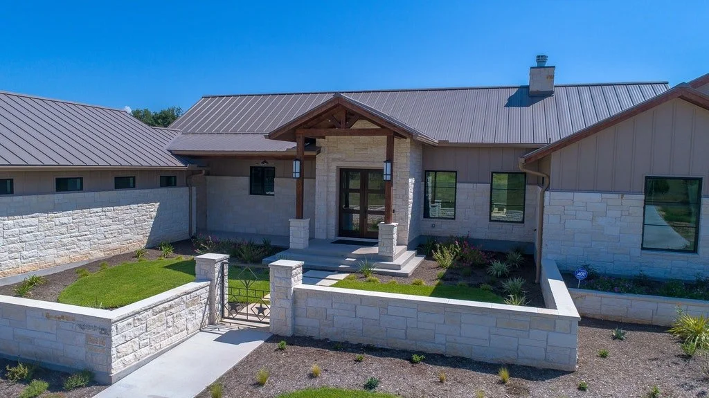 Modern house with stone and wood exterior, front steps, small landscaped yard, and a metal gate, under a clear blue sky.