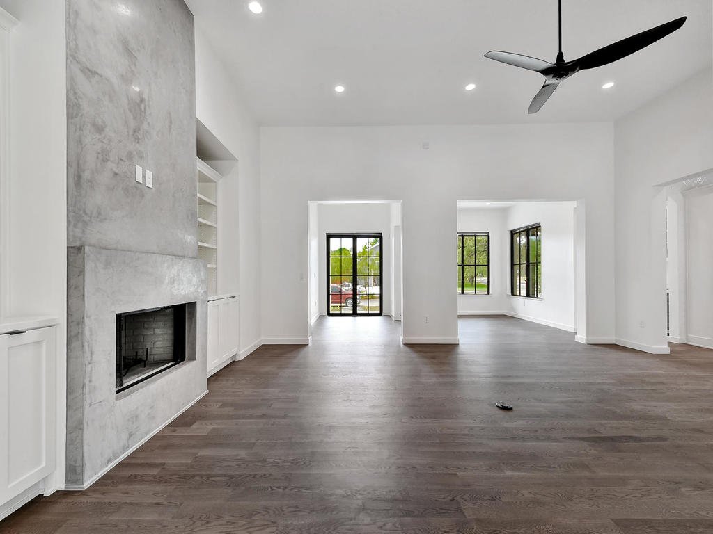Empty living room with hardwood floors, a modern fireplace, built-in shelves, large windows, a ceiling fan, and a view of the outdoors.