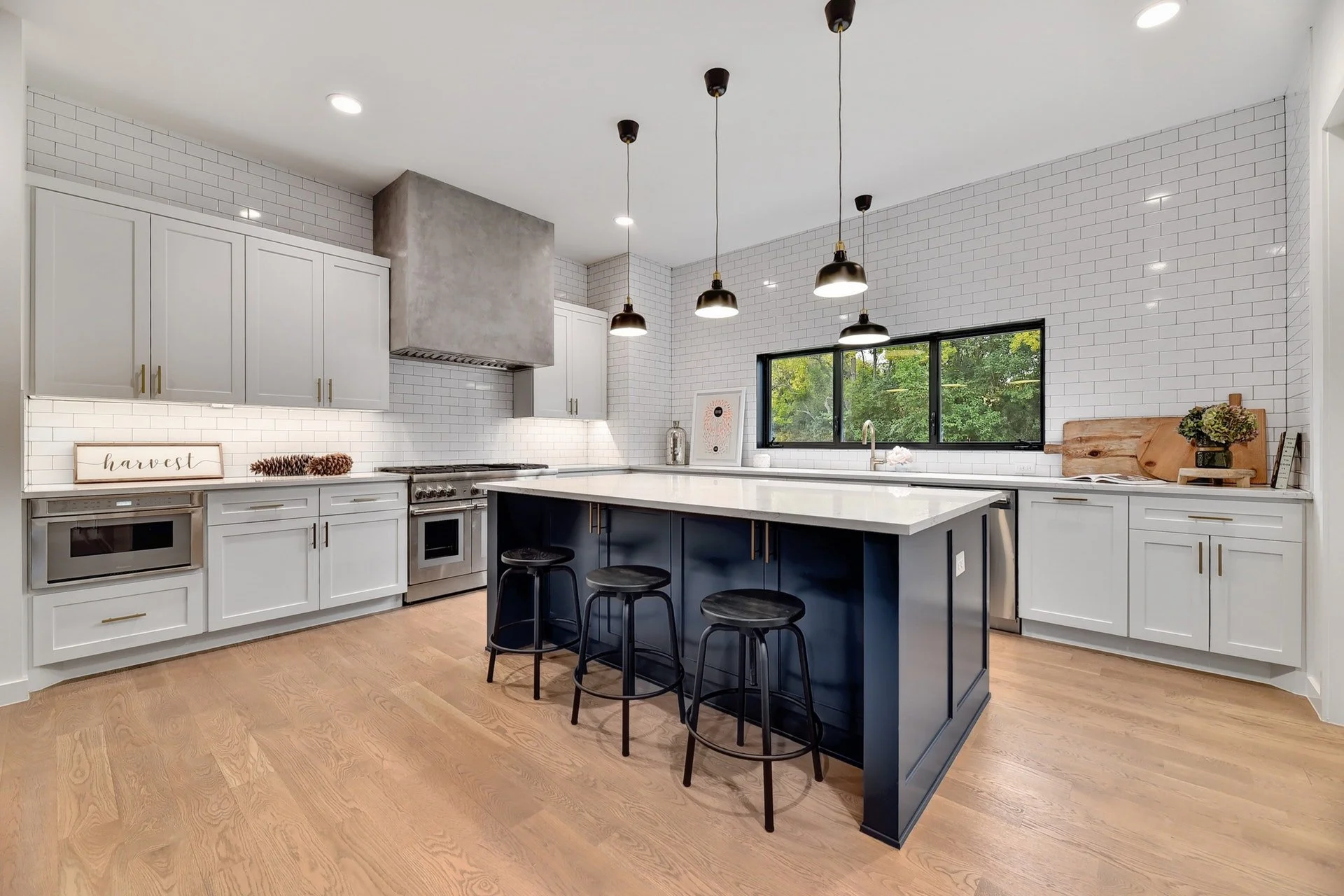Modern kitchen with white cabinets, a large island with a dark base, and black bar stools. White subway tile backsplash, stainless steel appliances, and a window showing green trees outside. Installing pendant lights and wooden accents.