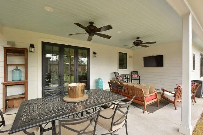 Covered outdoor patio with black metal dining table and chairs, a wooden sofa and armchairs with cushions, a wall-mounted TV, ceiling fans, and decor including a large blue vase and a table with a woven basket. Sliding glass doors lead to the interio
