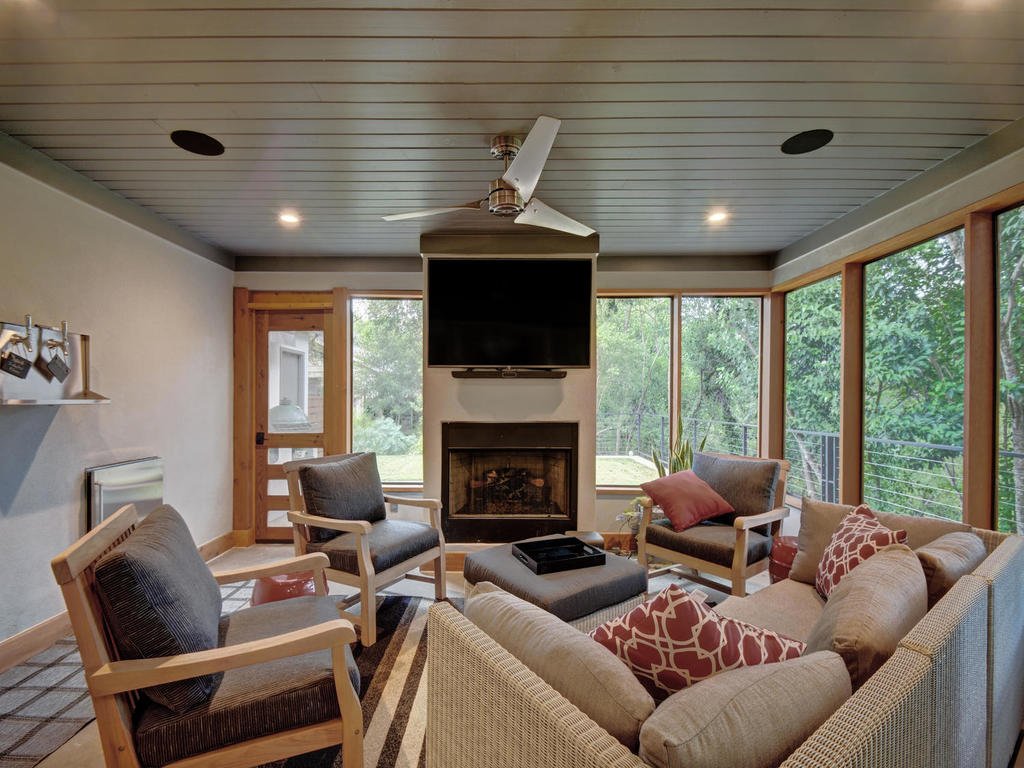 Living room with a fireplace, large windows showing greenery outside, several chairs and a sofa with decorative pillows, a wall-mounted TV above the fireplace, a ceiling fan, and a door leading outside.