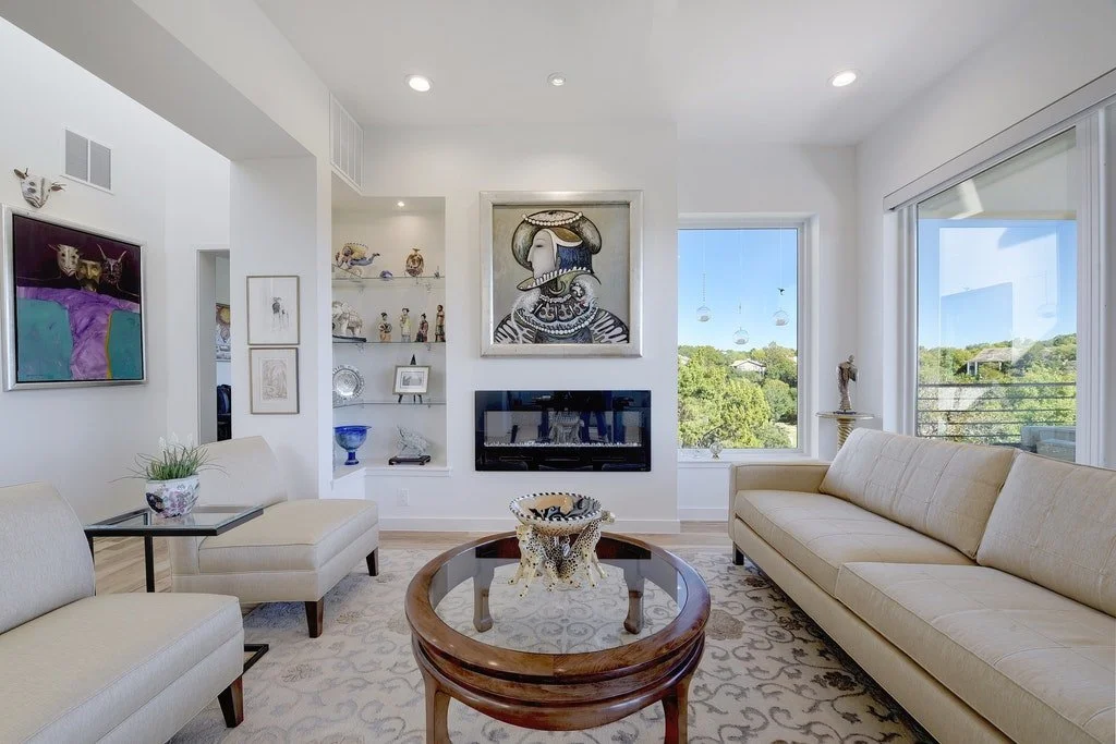Bright living room with beige sofas, glass top coffee table, wall-mounted fireplace, artwork, and large windows showing greenery outside.