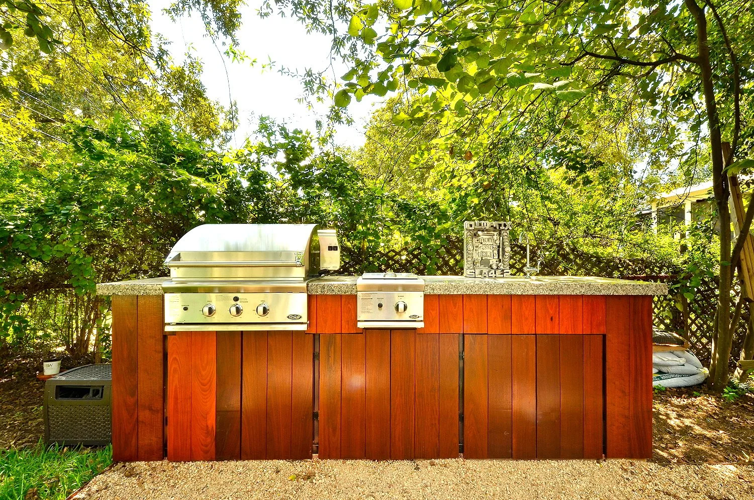 An outdoor kitchen setup with a granite countertop, a stainless steel grill, and a small stove, all housed within a wooden cabinet. There are lush green trees and shrubs in the background, creating a garden-like setting.