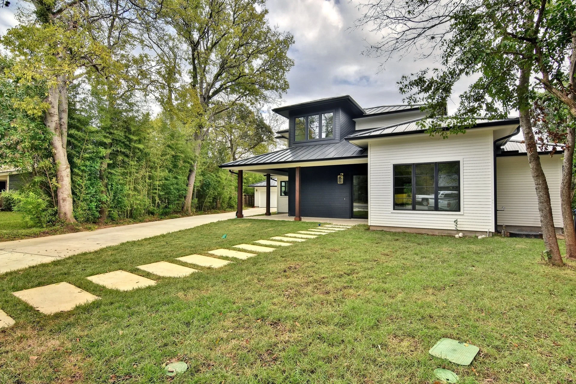 Modern two-story house with a white and dark gray exterior, large windows, a metal roof, and a front yard with grass, stepping stones, and trees on a cloudy day.