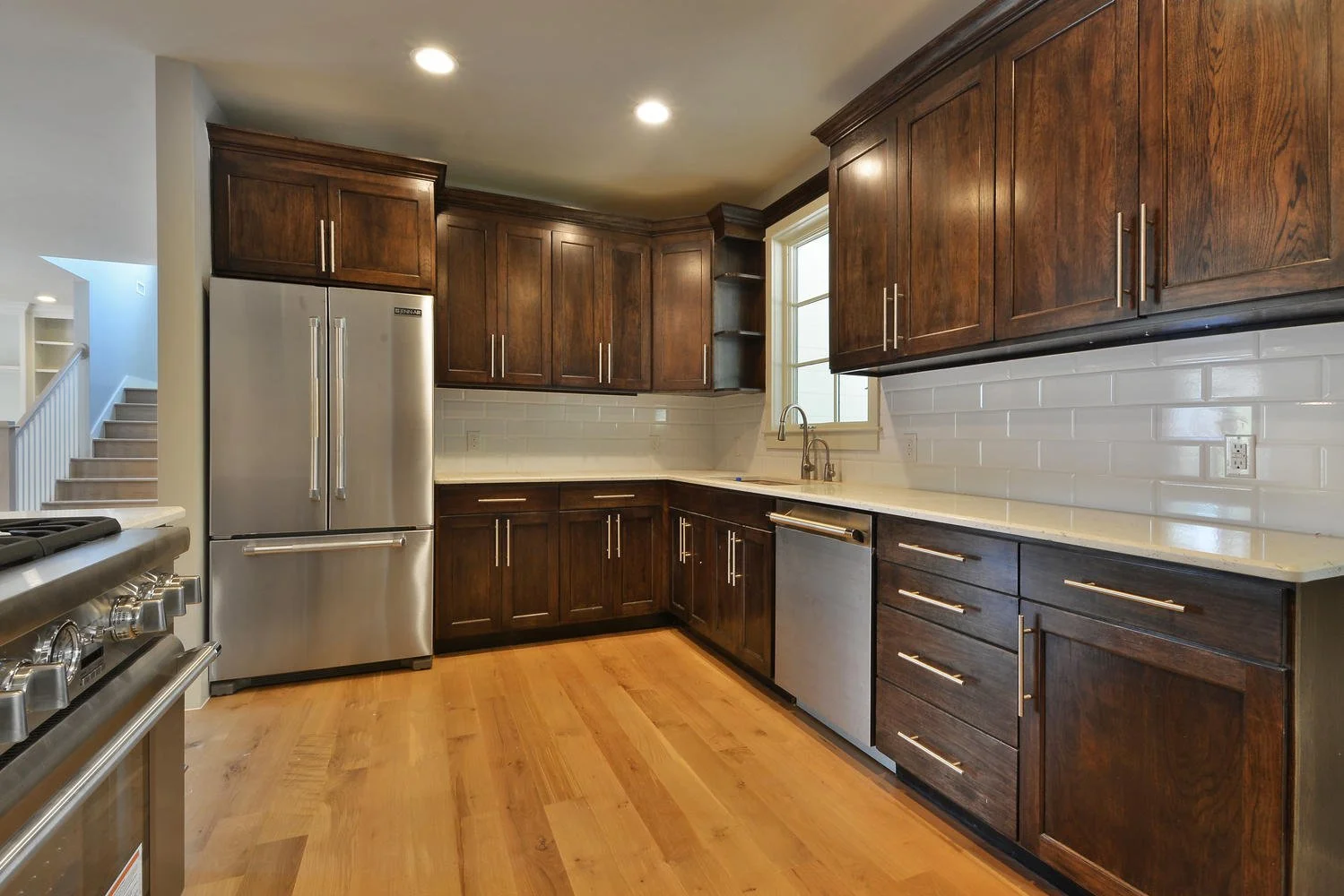 Modern kitchen with dark wood cabinets, stainless steel refrigerator, dishwasher, and gas stove, featuring a white subway tile backsplash and hardwood flooring.