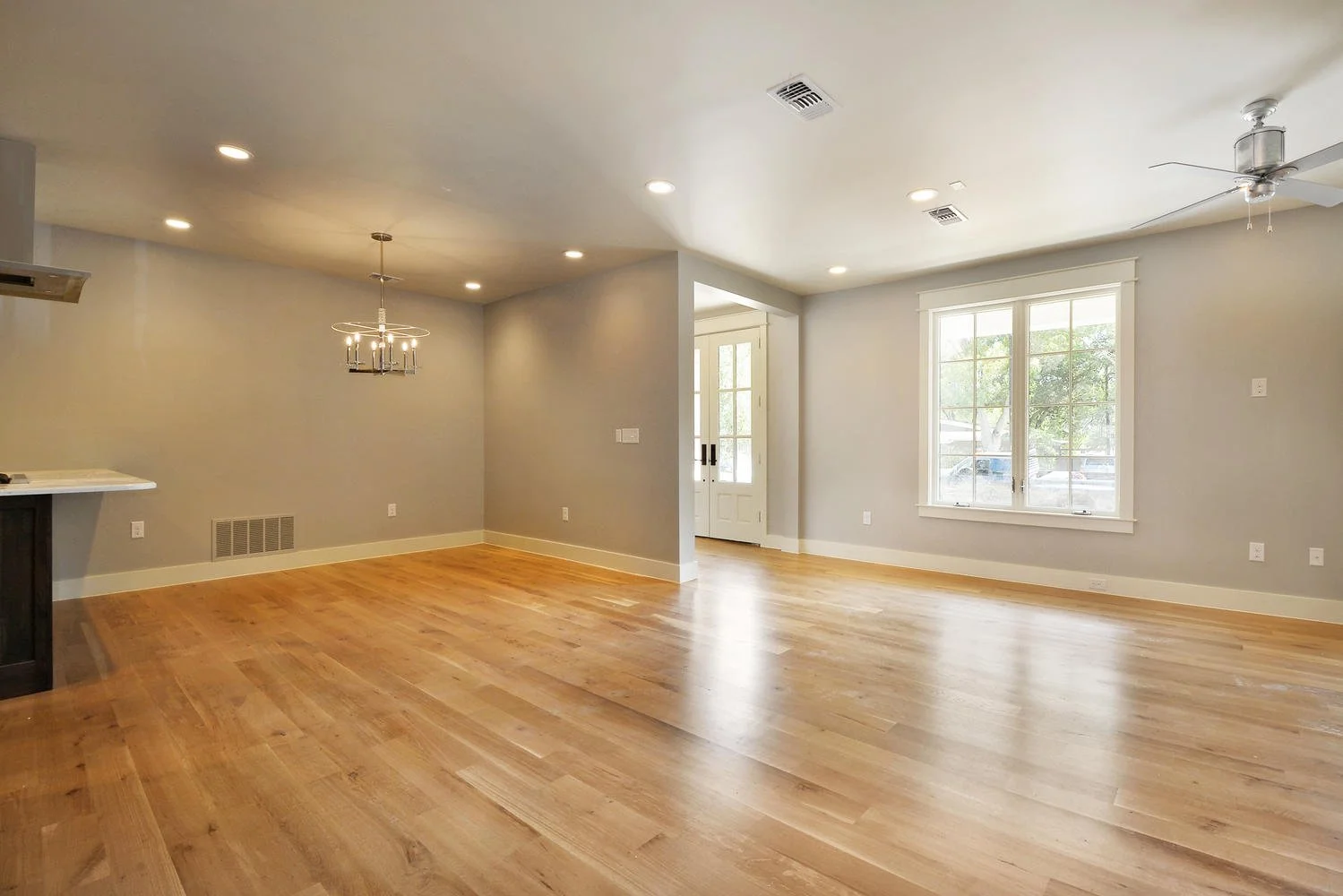 Empty living room with hardwood floors, large window, ceiling fan, recessed lighting, and front door.