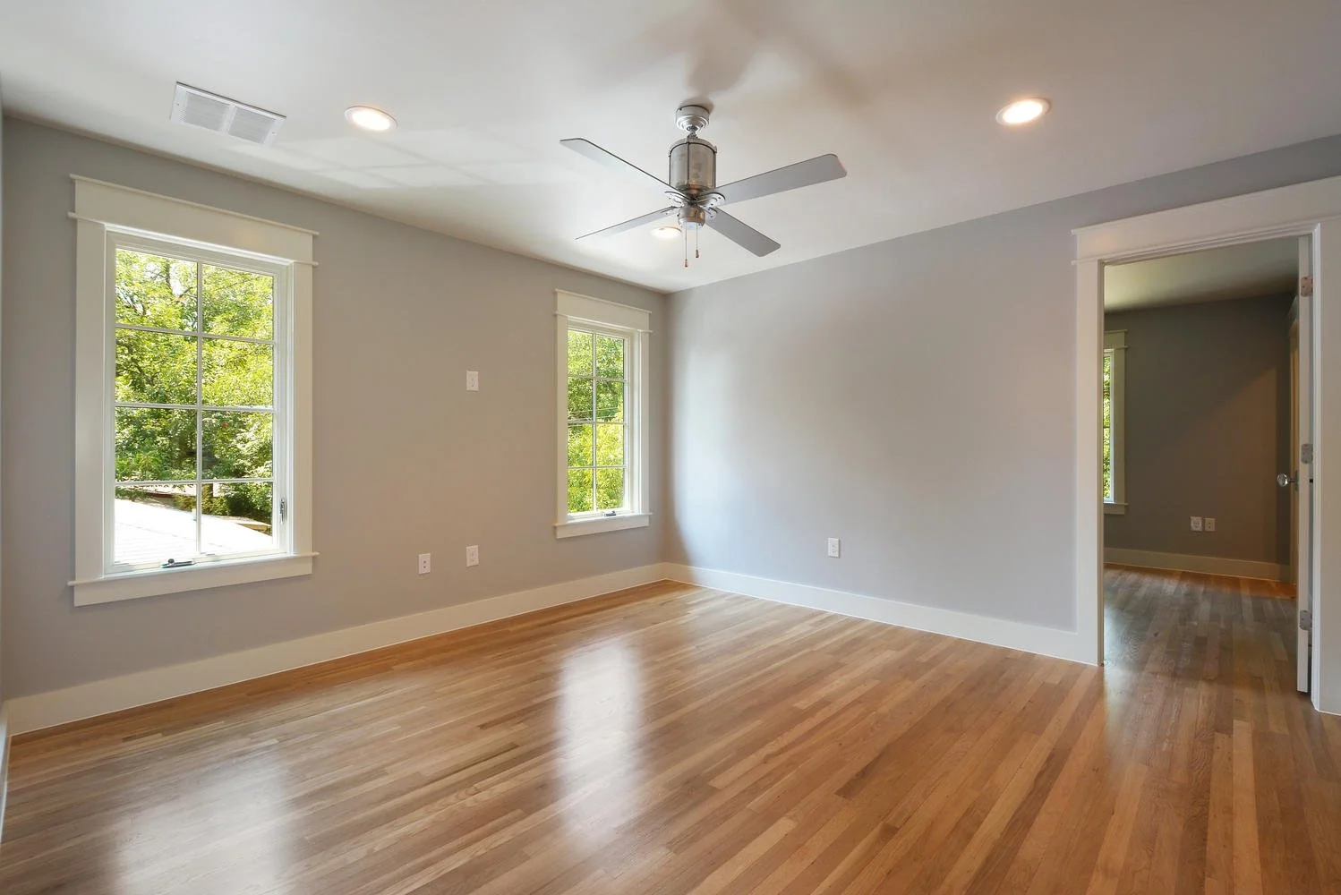 Empty room with light gray walls, hardwood flooring, two windows, white trim, ceiling fan, and an open doorway to another room.