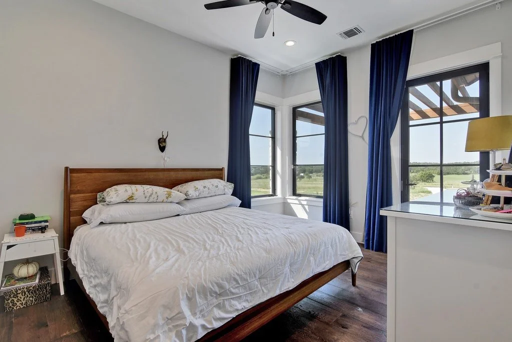 Bedroom with white walls, hardwood floor, wooden bed frame with white bedding, blue curtains, large windows showing outdoor scenery, white dresser, and a ceiling fan.