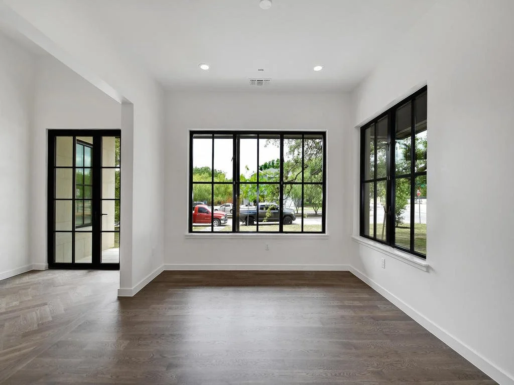 Empty room with white walls, large black-framed windows, and wood floors, overlooking a parking lot and greenery outside.