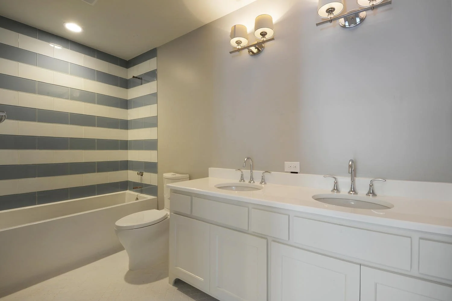 Modern bathroom with dual sinks, a bathtub with striped tile wall, a toilet, and a wall-mounted light fixture.