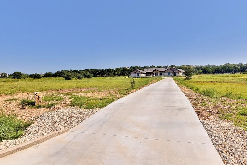 A paved driveway leading to a modern house in a grassy field under a clear blue sky.